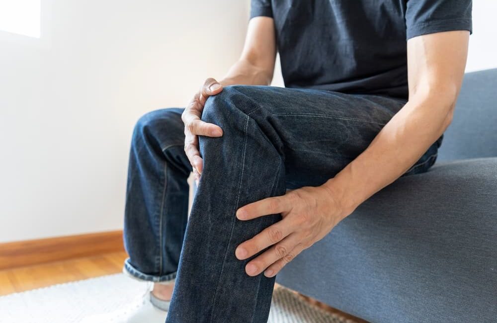 Close-up of a man's knee experiencing severe joint pain while sitting on a sofa.