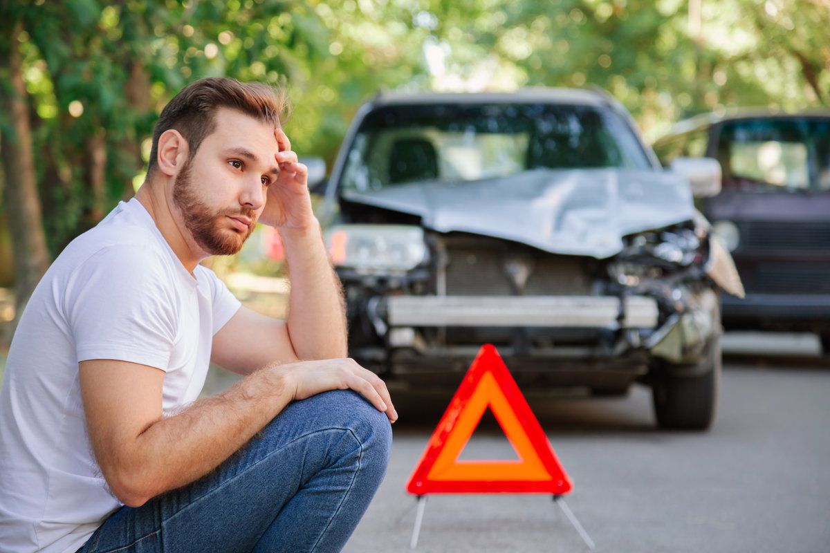 Portrait of upset man sitting on road near wrecked car after car accident with red triangle. Caucasian scary driver man sitting near his wrecked car on floor waiting for the insurance service.
